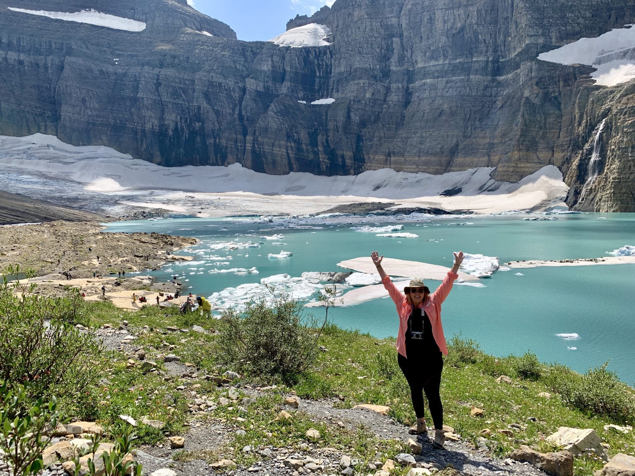 lady living in an RV standing in front of crystal blue Grinnell Glacier in Glacier National Park with gray jagged rocks in the background