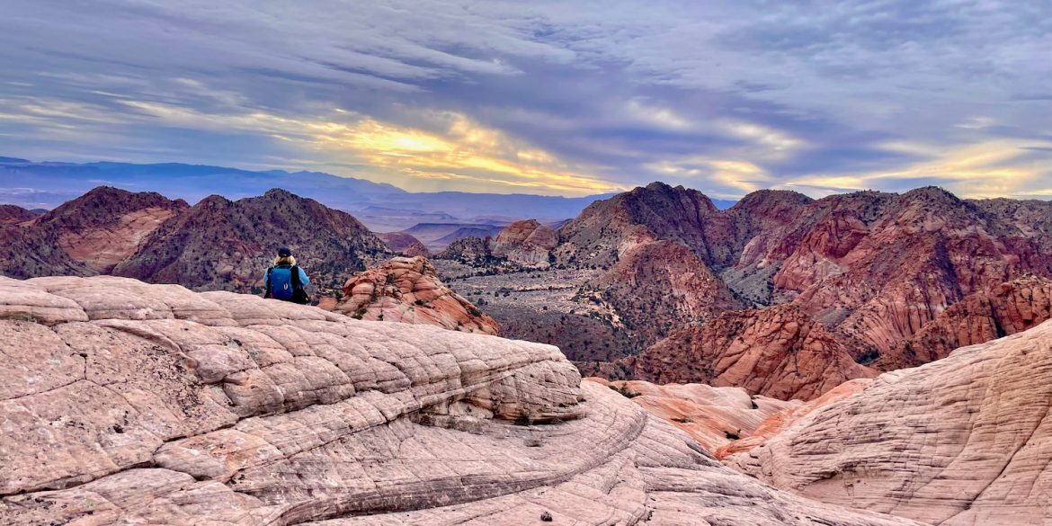 Woman living in an RV with backpack sitting with back shown gazing on sunset over the Candy Cliffs of Yant Flat.