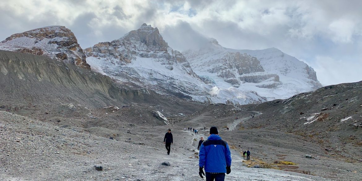 man living in an RV in blue parka walking towards Athabasca Glacier in Jasper National Park. Gray snow covered mountain surrounding.