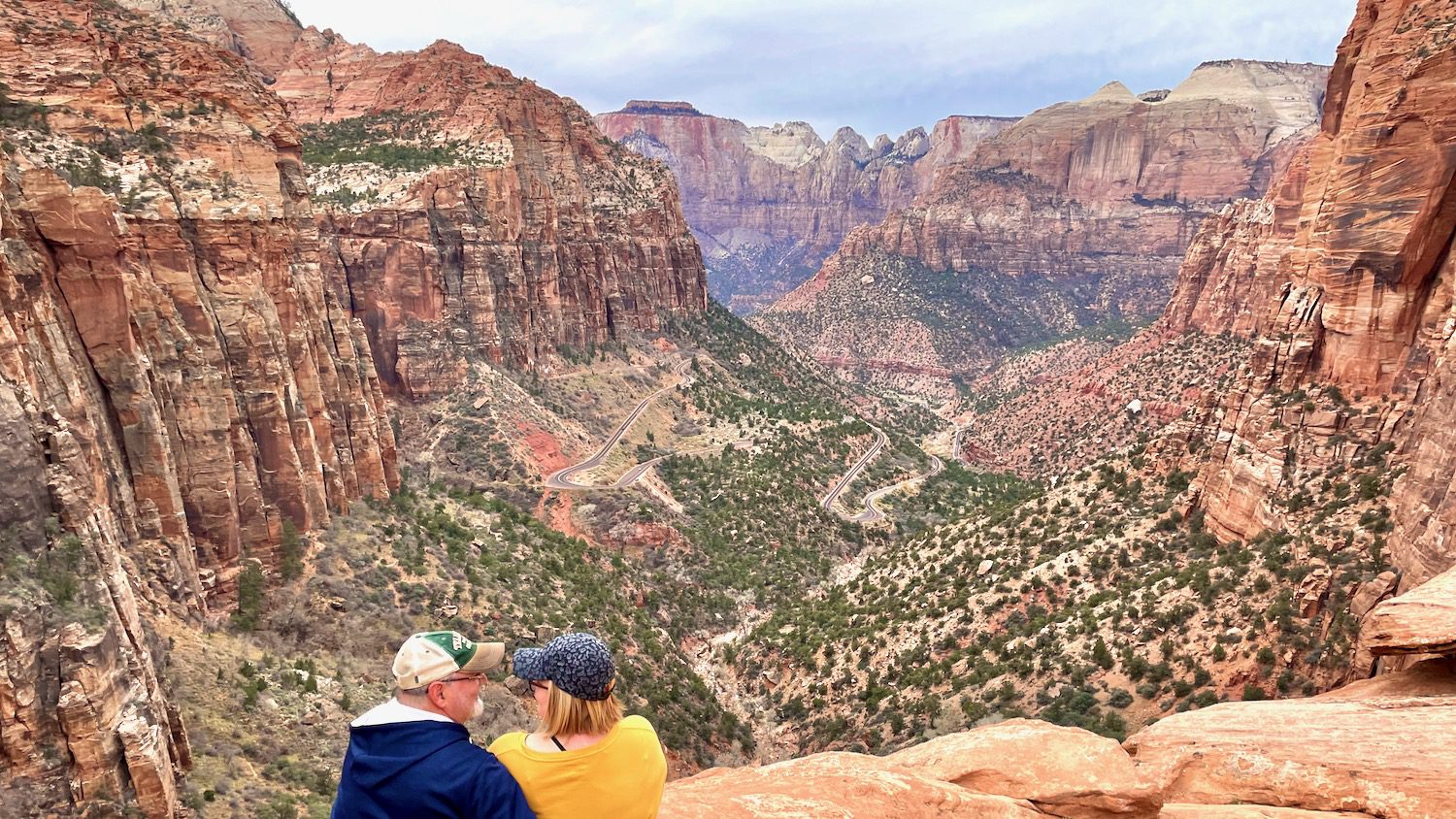 couple living in an RV looking at each other while sitting on the edge of a sandstone ledge overlooking a winding breathtaking mountain road in Zion National Park